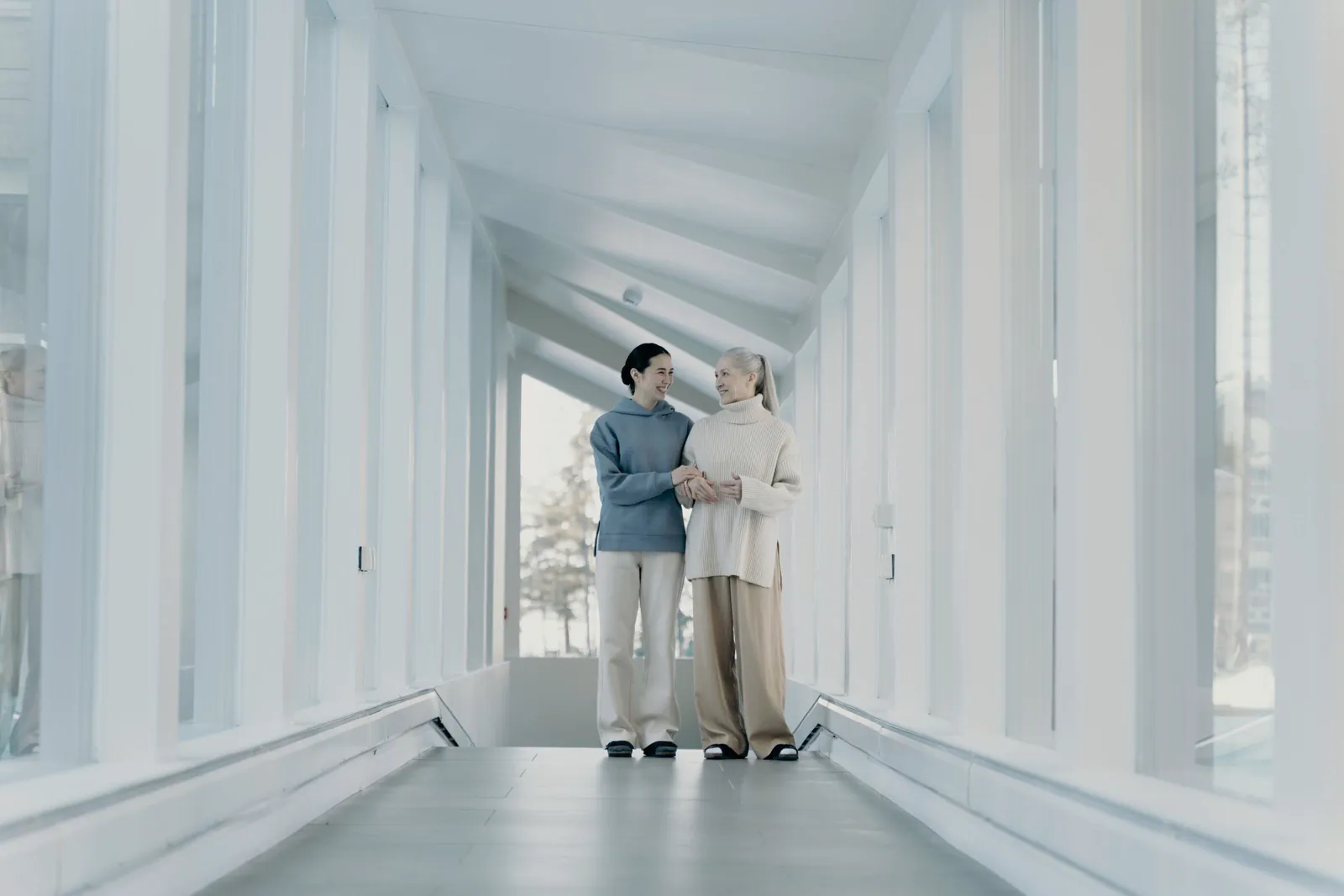 A younger woman walking arm in arm with an older woman along a bright glass corridor.
