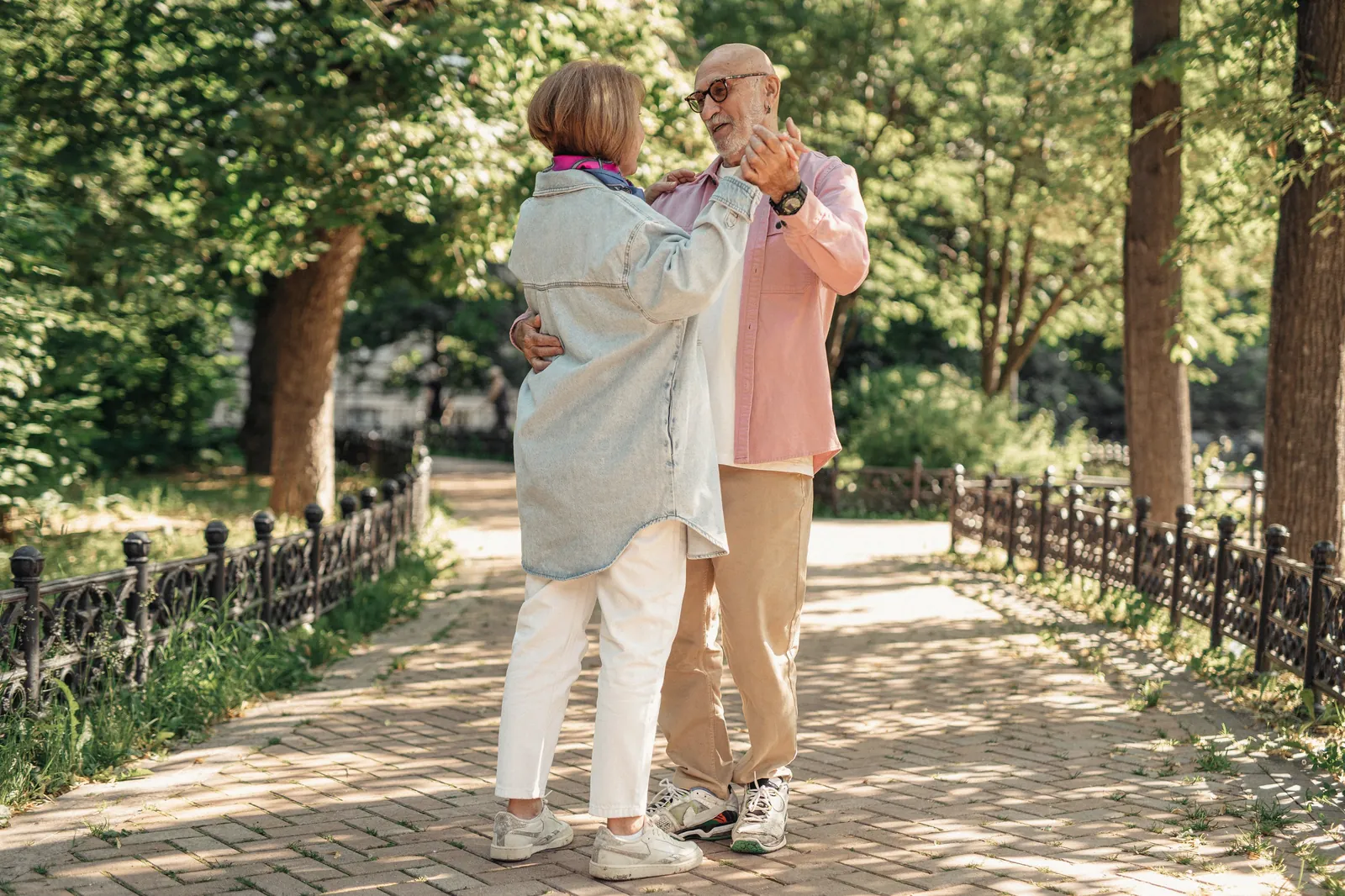 An older couple dancing together on a sunlit park path.